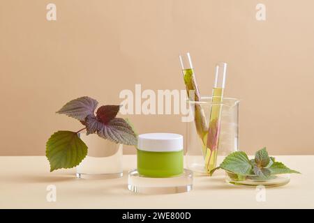 Pot vert avec étiquette vierge placée sur un podium transparent, deux tubes à essai de liquide jaune et quelques feuilles de beefsteak présentés. Produit de beauté naturel ext Banque D'Images