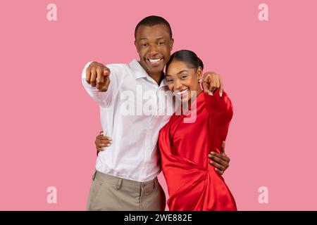Couple noir souriant pointant vers la caméra, homme en blanc, femme en rouge, fond rose Banque D'Images