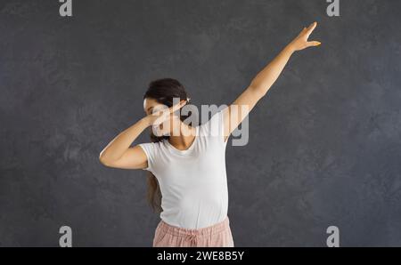 Joyeux drôle jeune femme caucasienne faisant geste de danse dab debout sur fond gris. Banque D'Images