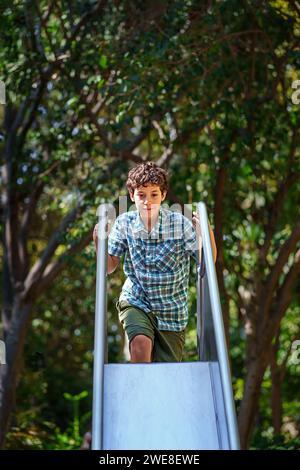 Jeune garçon glisse avec joie sur un toboggan de terrain de jeu, entouré de verdure par une journée ensoleillée, capturant l'essence du jeu d'enfance. Banque D'Images