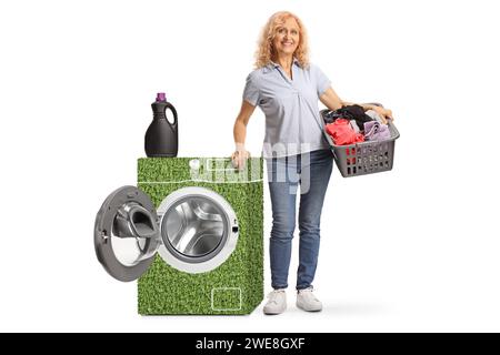 Portrait en longueur d'une femme tenant un panier à linge à côté d'une machine à laver à faible consommation d'énergie verte isolée sur fond blanc Banque D'Images