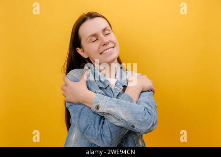 Portrait de gentille belle jeune femme debout avec les yeux fermés s'embrassant elle-même, étant égoïste, portant une veste en denim, posant isolé sur le jaune Banque D'Images