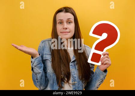 Portrait de jeune femme réfléchie confuse regardant la caméra, tenant le papier point d'interrogation rouge, pense aux tâches, portant une veste en denim, posant isola Banque D'Images