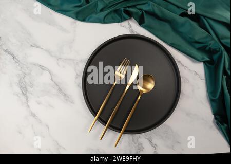 Vue de dessus de couverts dorés et assiette noire sur fond de marbre blanc. Tissu de soie vert, matériau satiné. Réglage de table de luxe, pose à plat, espace de copie. Banque D'Images