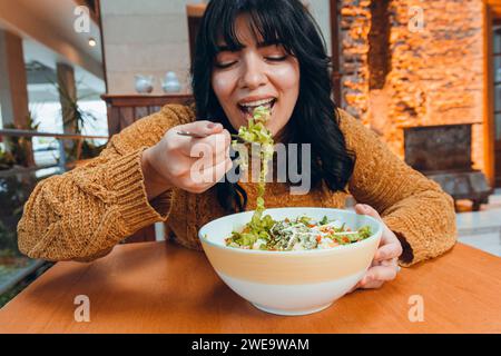 jeune femme vénézuélienne latine avec la bouche ouverte et les yeux fermés, appréciant et mangeant de la salade avec fourchette dans le restaurant, vue de face Banque D'Images