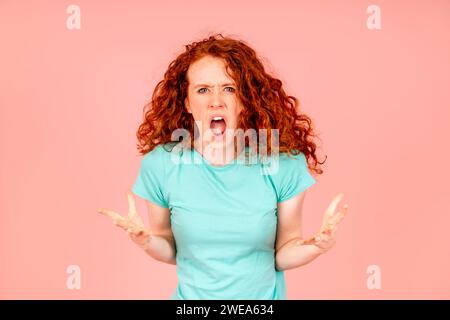 Jeune femme aux cheveux rouges en colère avec des cheveux bouclés ondulés avec des oreilles fermées en t-shirt vert crie et flippe sur fond rose J'en ai fini avec tout le monde Banque D'Images