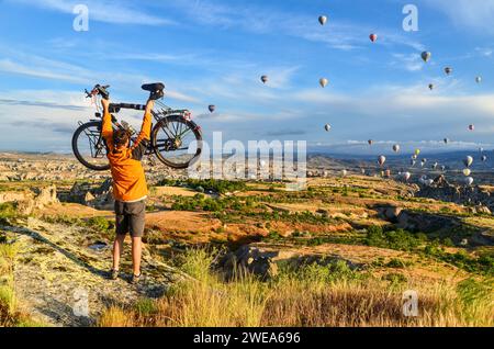 Homme soulevant son vélo de tourisme en Cappadoce, devant les ballons à air levant dans les premières heures d'une journée ensoleillée Banque D'Images