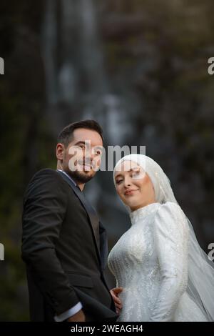 Prise de vue à faible angle de jeunes mariés heureux debout devant une cascade souriant et posant à l'extérieur Banque D'Images