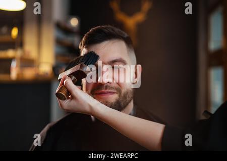 Portrait d'homme barbu dans la chaise de salon de coiffure appréciant le traitement de coiffure tout en nettoyant doucement le visage du bras de barbier féminin à l'aide de la brosse de cou. Éclairage tamisé moderne Banque D'Images