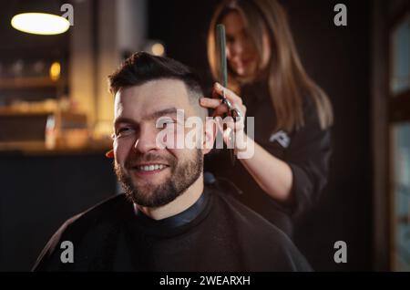 Portrait d'homme barbu souriant tout en coupe coiffure dans salon de coiffure par jeune femme coiffeuse. salon de coiffure moderne de style noir et sombre Banque D'Images