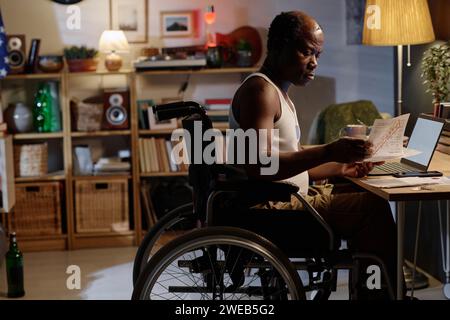 Homme afro-américain handicapé assis à son bureau à la maison et regardant à travers des papiers Banque D'Images