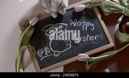 Un tableau noir avec un lapin de pâques et Joyeuses Pâques dessus. Tulipes roses. Rose, fond violet. Concept de vacances. Banque D'Images