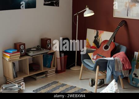 Nouvelle guitare acoustique debout sur fauteuil avec pile de vêtements devant le tourne-disque entouré de livres et de deux haut-parleurs sonores Banque D'Images