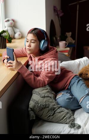 Mignonne adolescente dans des écouteurs se relaxant sur le lit par le rebord de la fenêtre et regardant l'écran du smartphone tout en regardant la vidéo en ligne Banque D'Images