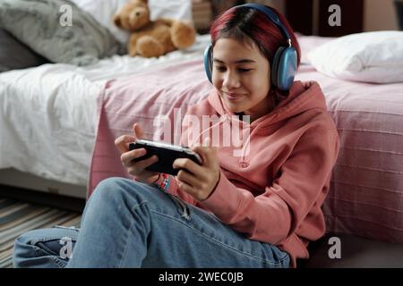 Heureuse adolescente avec smartphone dans GamePad assis sur le sol par son lit et regardant l'écran tout en jouant au jeu vidéo Banque D'Images