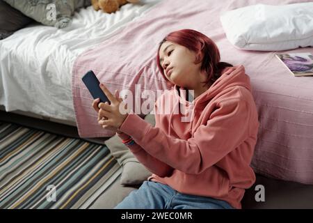 Adolescente en sweat à capuche rose assise sur le sol par le lit et regardant l'écran du smartphone tout en communiquant dans le chat vidéo Banque D'Images