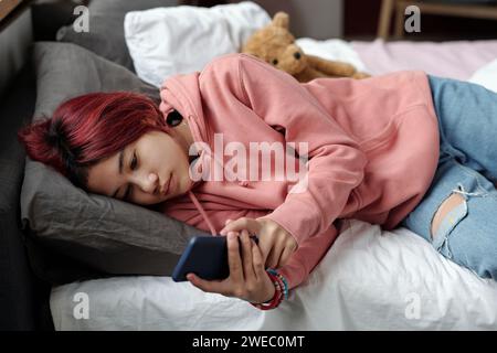 Joli adolescent en sweat à capuche rose se relaxant sur le lit et regardant l'écran du smartphone tout en regardant la vidéo sur Internet à loisir Banque D'Images