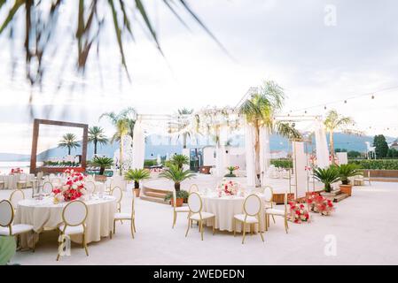 Des tables festives avec des bouquets colorés de fleurs se dressent autour d'une scène ronde sur le bord de la mer sur fond de montagnes Banque D'Images