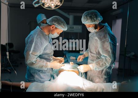 Chirurgiens professionnels ciblés et infirmière en uniforme au travail dans la salle d'opération, effectuant une opération de transplantation cardiaque sous des lampes brillantes. Banque D'Images