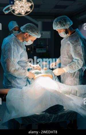 Portrait vertical de chirurgiens masculins concentrés avec une assistante féminine suture la peau de l'estomac pendant la chirurgie avec des points de suture nets après élimination de l'hygroma Banque D'Images