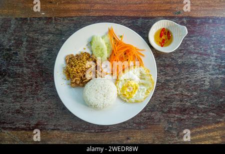 Nourriture thaïlandaise mélanger porc frit avec ail et poivrons thaïlandais sur le dessus ail frit croustillant servi avec riz, œuf frit et concombre de tomate assiette blanche sculptée à Banque D'Images