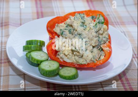 Salade d'œufs Herby avec poulet râpé et épinards hachés farcis dans un poivron rouge avec des mini concombres sur une assiette blanche sur une nappe à carreaux. Banque D'Images