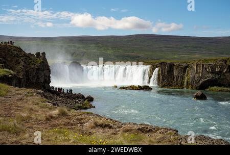 De minuscules touristes voient la majestueuse cascade Godafoss sur la rivière Skjalfandafljot dans le nord de l'Islande avec une belle colline verdoyante en arrière-plan Banque D'Images