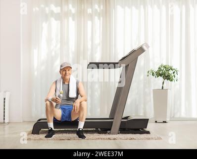 Homme âgé reposant sur un tapis roulant à la maison avec une bouteille d'eau Banque D'Images