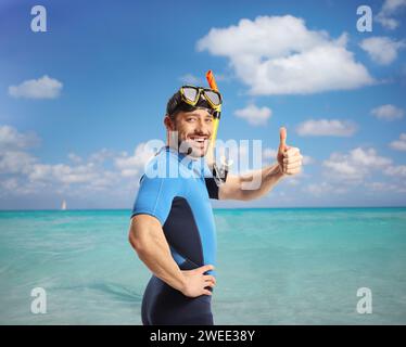 Jeune homme en combinaison de plongée et masque faisant des gestes de pouces vers le haut à l'intérieur de la mer bleue Banque D'Images