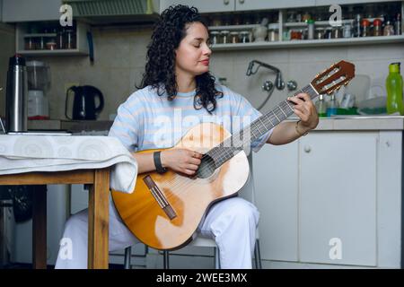 jeune femme vénézuélienne latine assise à pratiquer la guitare acoustique à la maison, jouer des accords faisant de la musique, pratiquer des chansons. vue avant. Banque D'Images