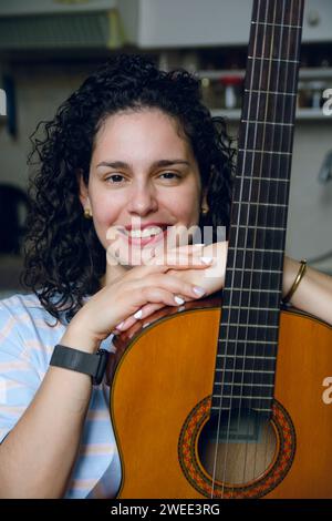 portrait vertical de jeune femme latine avec des bigoudis, assise à la maison calme heureuse et souriante avec sa guitare regardant la caméra Banque D'Images