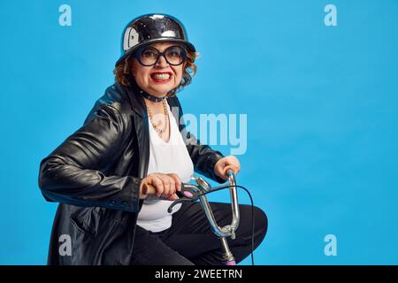 Portrait de vêtements en cuir vintage habillés et dans le casque chevauchant le vélo des enfants et souriant regardant la caméra sur fond bleu. Banque D'Images