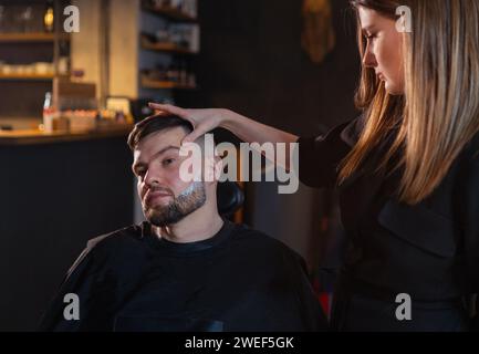 Une jeune femme coiffeuse aux cheveux longs apprécie les bords des poils après avoir rasé le visage barbu avec des restes de mousse crème. Appareil pour soins de la peau et des cheveux Banque D'Images