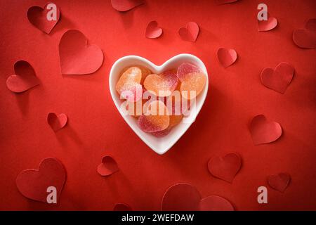 Fond d'amour conceptuel avec récipient en céramique blanche avec des bonbons en forme de coeur sur fond rouge avec des découpes en forme de coeur rouge. Vue de dessus. Banque D'Images