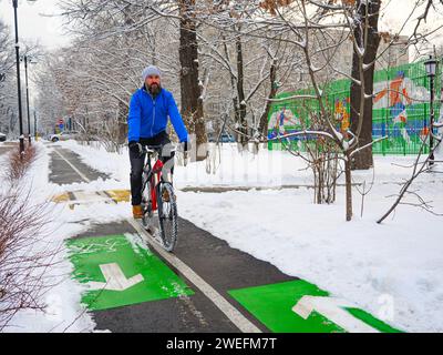 Un homme barbu fait du vélo le long d'une piste cyclable dans un parc en hiver. Mode de vie actif en hiver. Homme dans une veste bleue sur un vélo rouge Banque D'Images