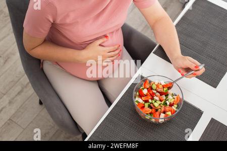 Une fille enceinte avec un gros ventre est assise dans la cuisine et mange une salade de vitamines végétales. Une bonne nutrition pendant la grossesse. Banque D'Images