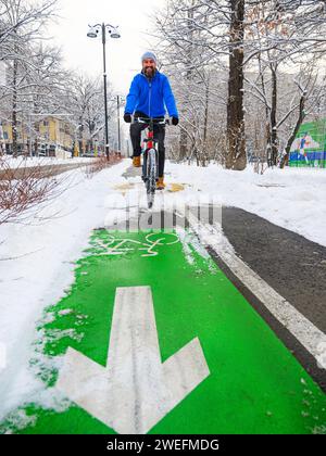 Un homme souriant fait du vélo le long d'une piste cyclable en hiver. Mode de vie actif en hiver. Homme barbu dans une veste bleue sur un vélo rouge Banque D'Images