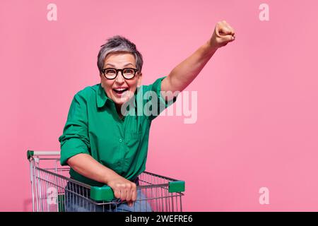 Photo de la femme satisfaite overjoyed porter une chemise verte assis dans le panier levant le poing hurlant isolé sur fond de couleur rose Banque D'Images