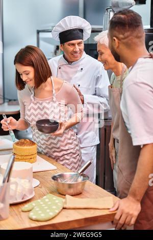 jeune femme joyeuse brossant le gâteau avec du sirop tandis que ses amis divers et chef parlent activement Banque D'Images