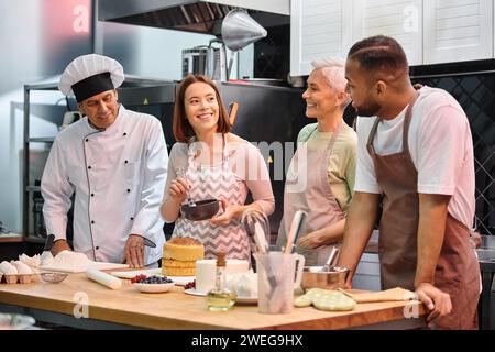 joyeuse jeune femme brossant le gâteau avec du sirop et souriant à ses amis divers à côté du chef mature Banque D'Images