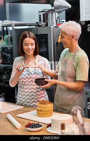 jeune femme donnant une brosse en silicone à son ami joyeux mature pour l'utiliser sur le gâteau, les cours de cuisine Banque D'Images