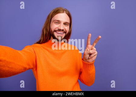 Autoportrait de gars hipster cheveux blonds longs portant un pull orange fait signe v envoyant des salutations isolé sur fond de couleur violette Banque D'Images