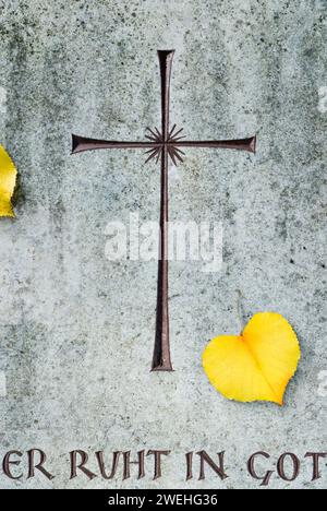 Une feuille en forme de cœur d'un tilleul d'hiver, feuille d'automne, repose sur une dalle de pierre tombale avec une croix et l'inscription il repose en Dieu Banque D'Images