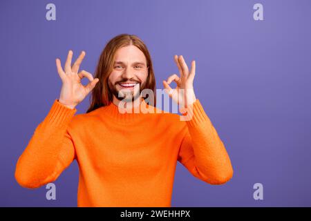 Portrait de jeune heureux homme de cheveux blonds pacifiques hippie portant un pull orange montrant double okey isolé sur fond de couleur violette Banque D'Images