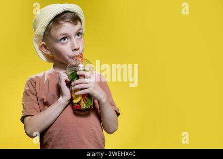 Garçon mignon au panama buvant un cocktail mojito de tasse en plastique sur fond de studio jaune. Banque D'Images