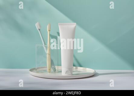 Vue de face du tube en plastique blanc sans étiquette, brosses à dents dans une tasse en verre sur plateau rond, sur fond bleu. Scène de maquette pour nettoyage cosmétique du visage Banque D'Images