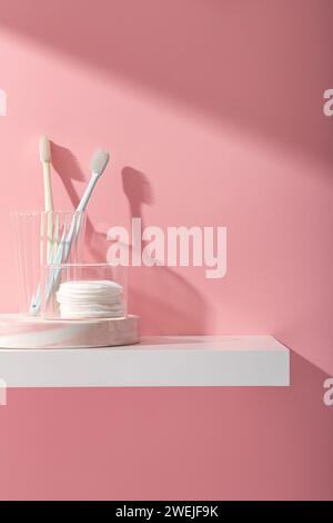 Scène publicitaire pour les produits de soins bucco-dentaires et de nettoyage avec des brosses à dents dans la tasse en verre et enlever le coton de maquillage sur le piédestal, sur fond rose. Espace f Banque D'Images