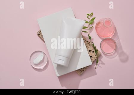 Une maquette de tube en plastique blanc pour cosmétique placée sur un podium blanc et des briques empilées. Verrerie de laboratoire contenant du liquide rose et de la texture crème sur podiu Banque D'Images