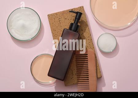 Une bouteille ambrée sur briques brunes, peigne en bois et boîtes de Pétri contenant du liquide et de la mousse sur fond rose. Emballage de produits de beauté pour soins capillaires. Production Banque D'Images