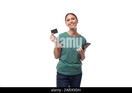 Femme brune européenne mince de 30 ans avec une queue de cheval habillée d'un t-shirt vert et un Jean tient une carte en plastique et un téléphone. Banque D'Images